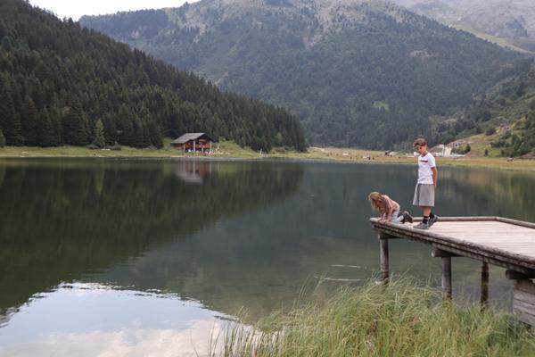 Two children are on a wooden pier, one intently looking into the clear mountain lake while the other stands nearby